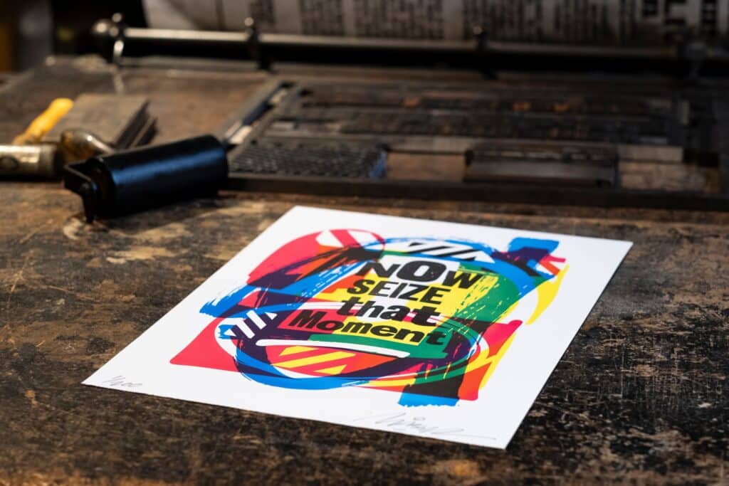 The print Parallels by Maser, with the text 'Now seize that moment', on the bed of a Wharfdale Printing Press