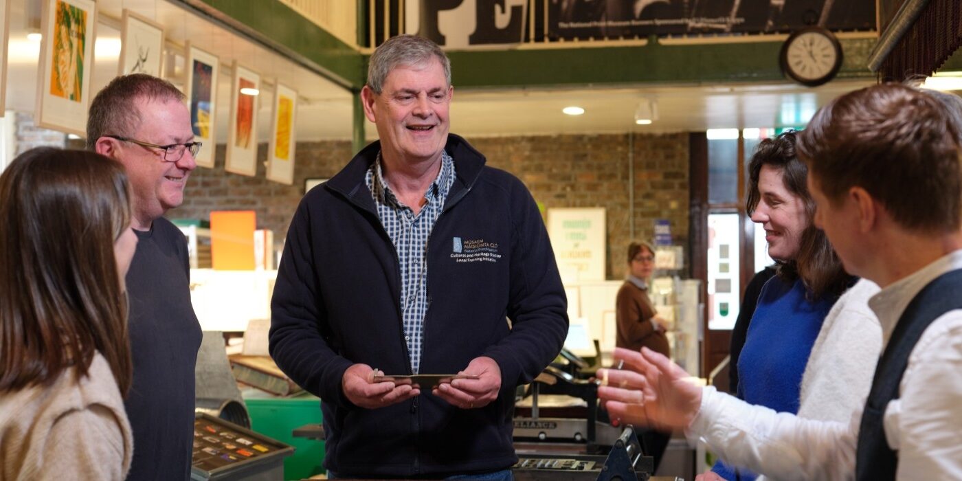A tour guide holds a composing stick and speaks to tour attendees
