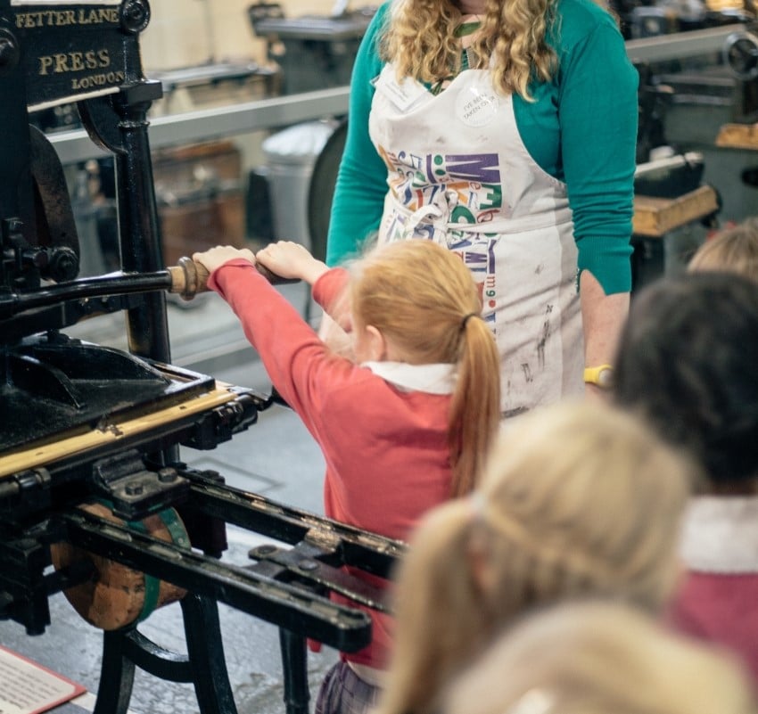 A tour guide supervises a student to pull the handle on an Albion printing press