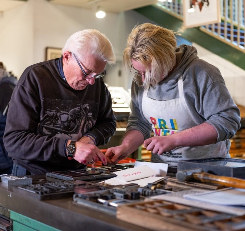 A printer and trainee adjust a forme at a composing stone