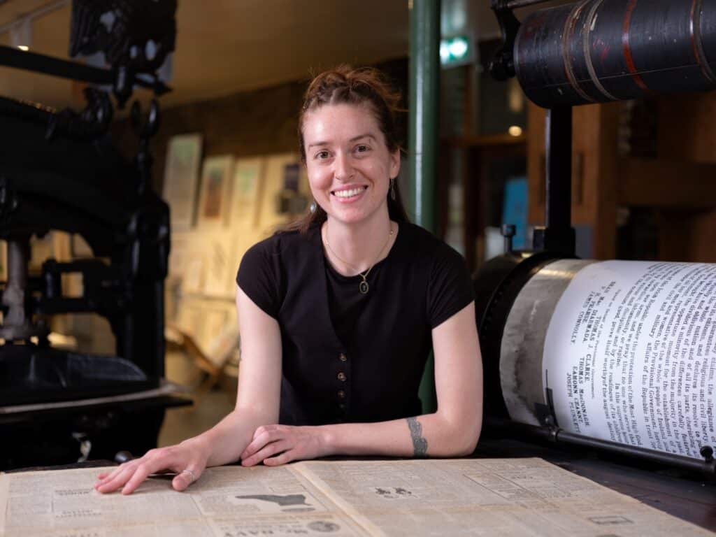 Work placement student Mazie stands at the Wharfdale stop-cylinder press with some Newspapers from the Museum's archive