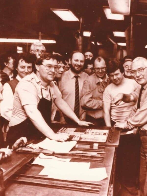 Printers and journalists gather around a stone with a newspaper forme on top