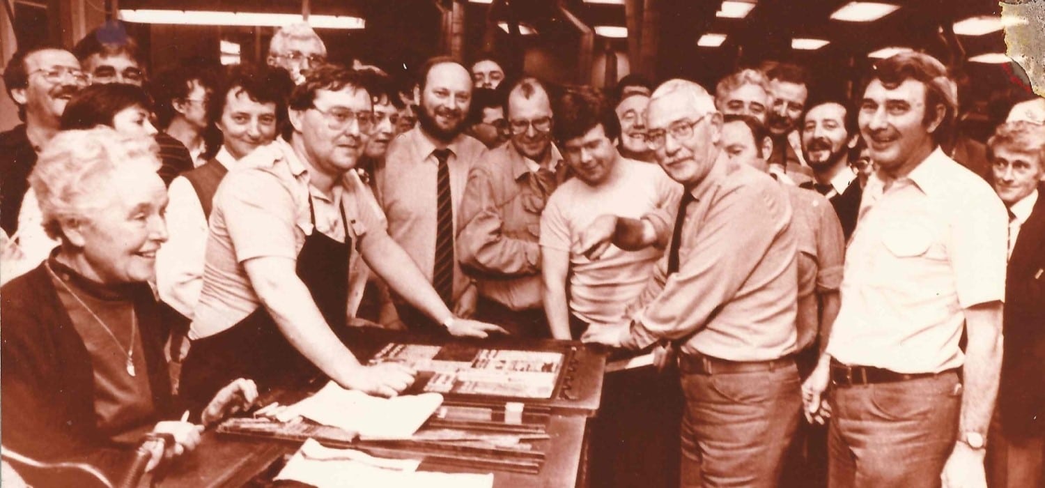 Printers and journalists gather around a stone with a newspaper forme on top