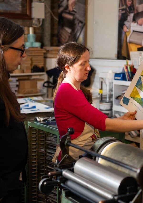 Two designers look at printers' proofs behind held up in front of a printing press.