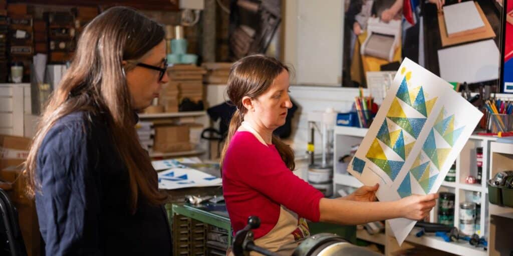 Two designers look at printers' proofs behind held up in front of a printing press.