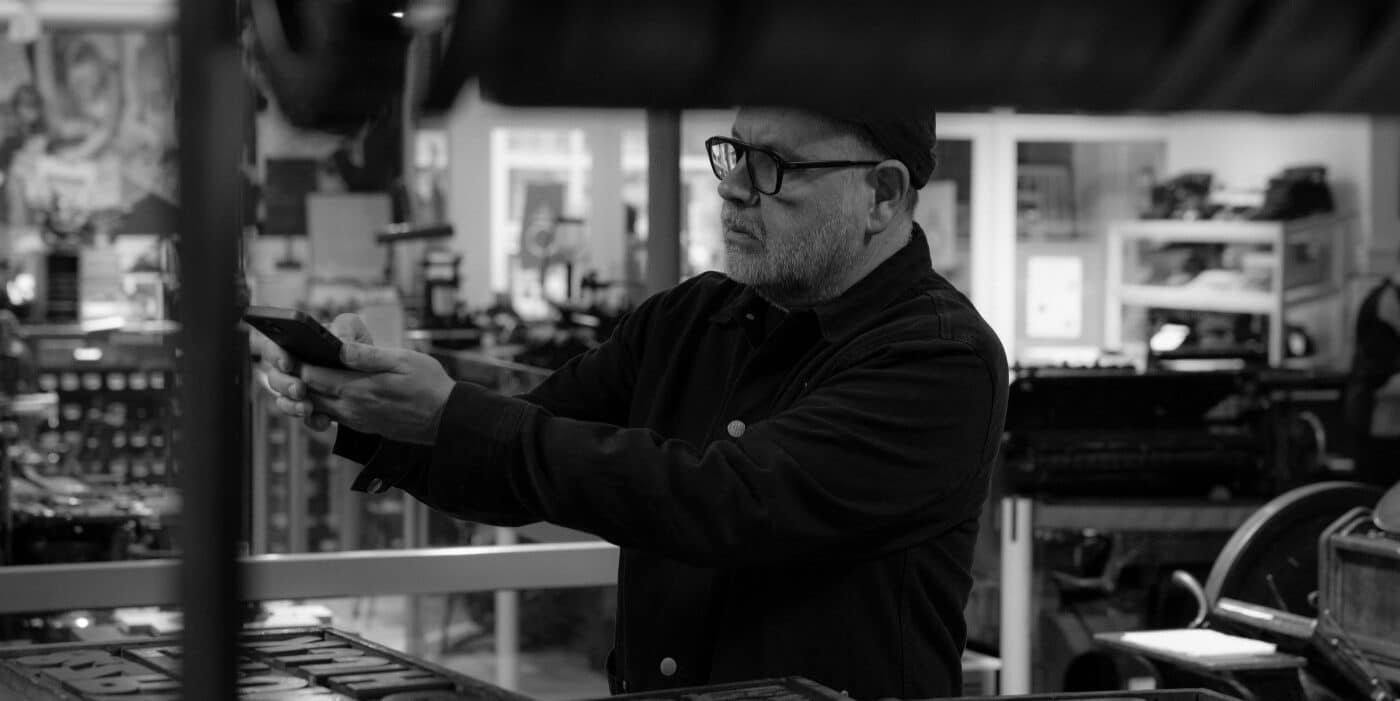 The artist Anthony Burrill holds up and looks at a piece of wooden type next to a printing press.