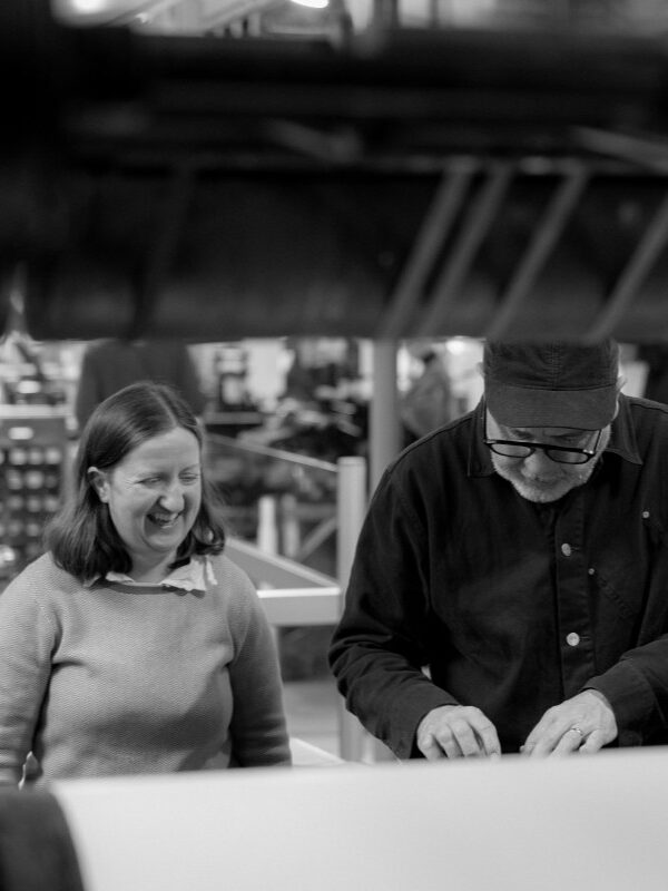 Mary Plunkett and Anthony Burrill look down at items laid out on a Wharfedale printing press.