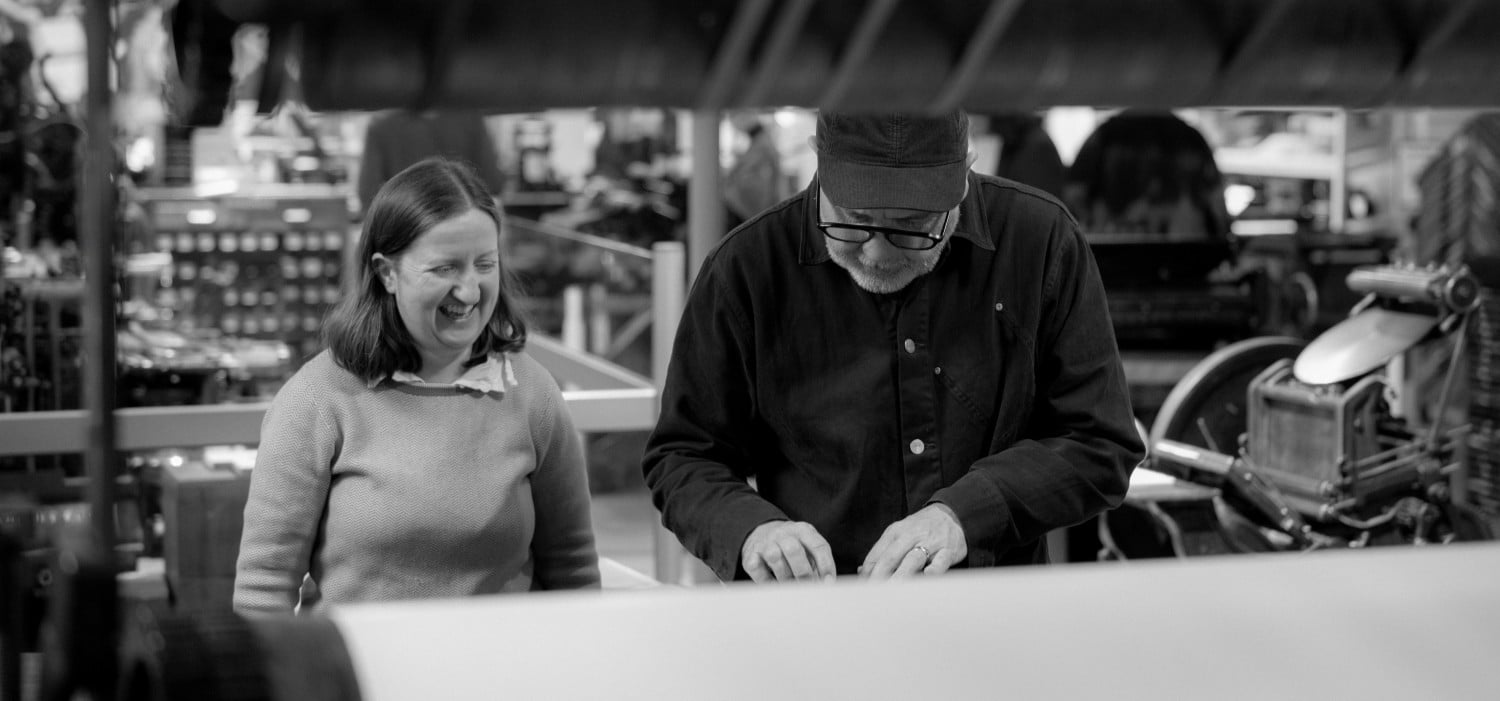 Mary Plunkett and Anthony Burrill look down at items laid out on a Wharfedale printing press.