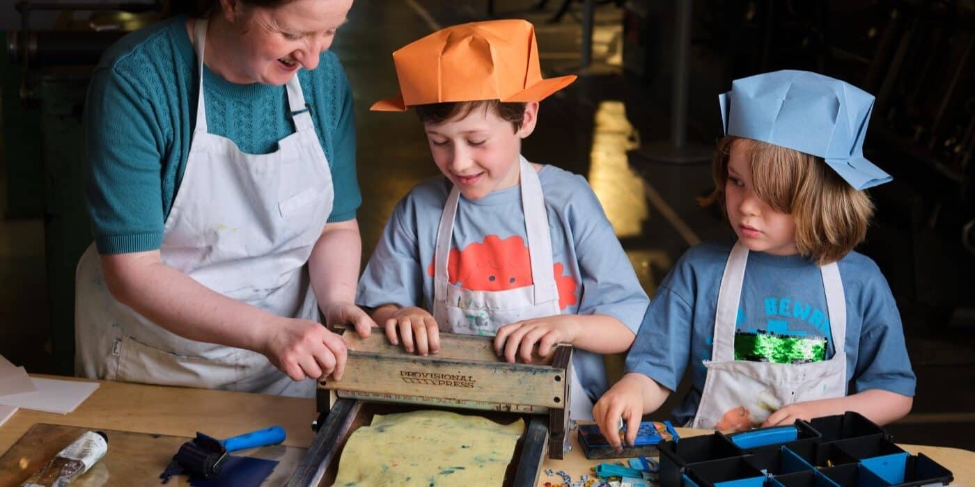 A woman helps two young boys to print using LEGO on a small printing press.