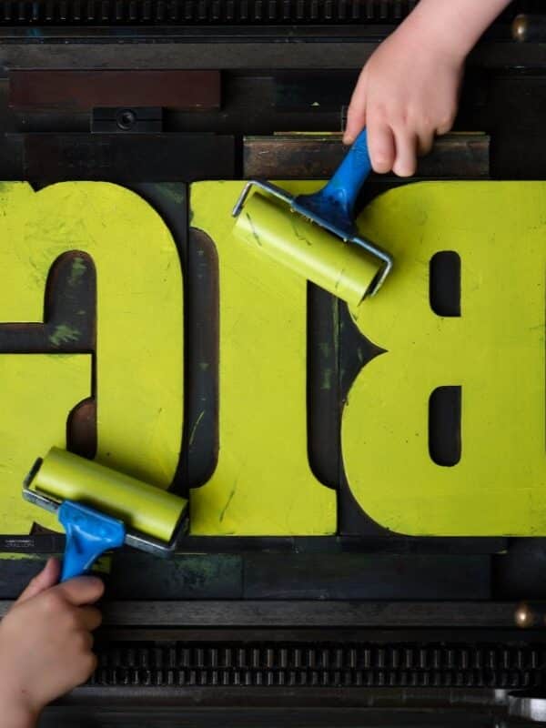 The word BIG is set on a printing press and in the process of being inked in yellow by two hand ink rollers.