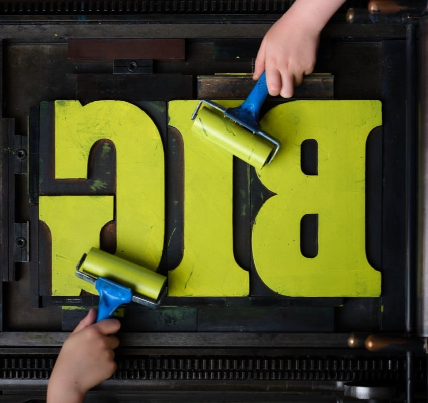 The word BIG is set on a printing press and in the process of being inked in yellow by two hand ink rollers.