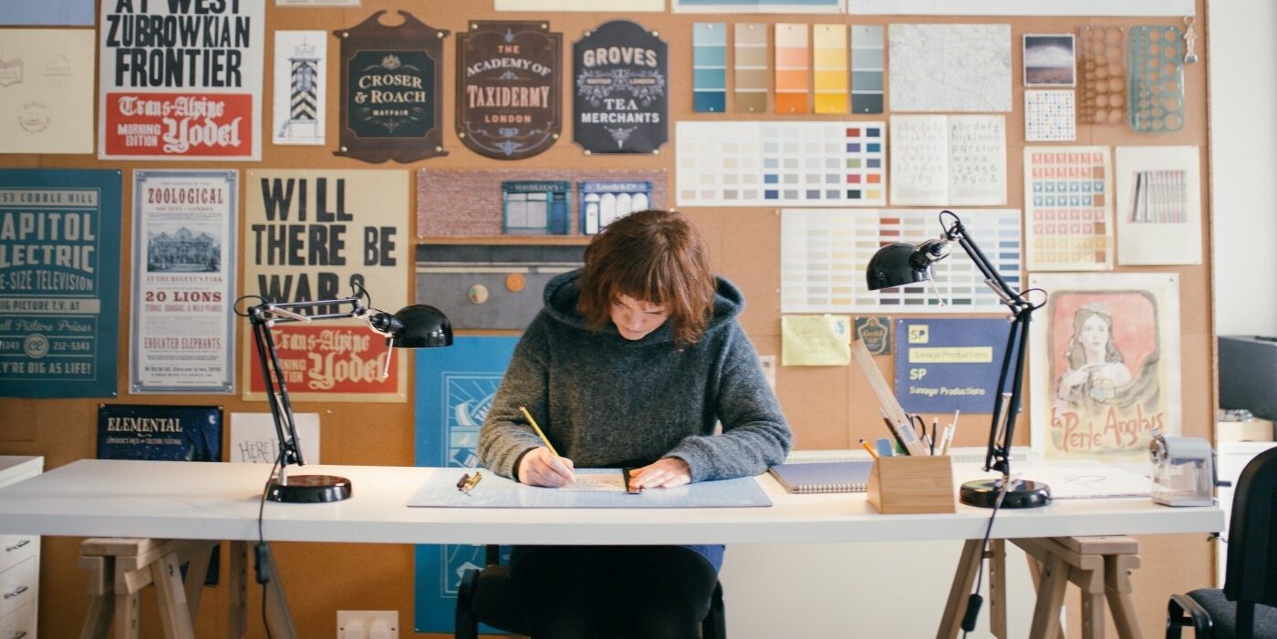 A photograph of artist Annie Atkins looking down as she works at a desk.