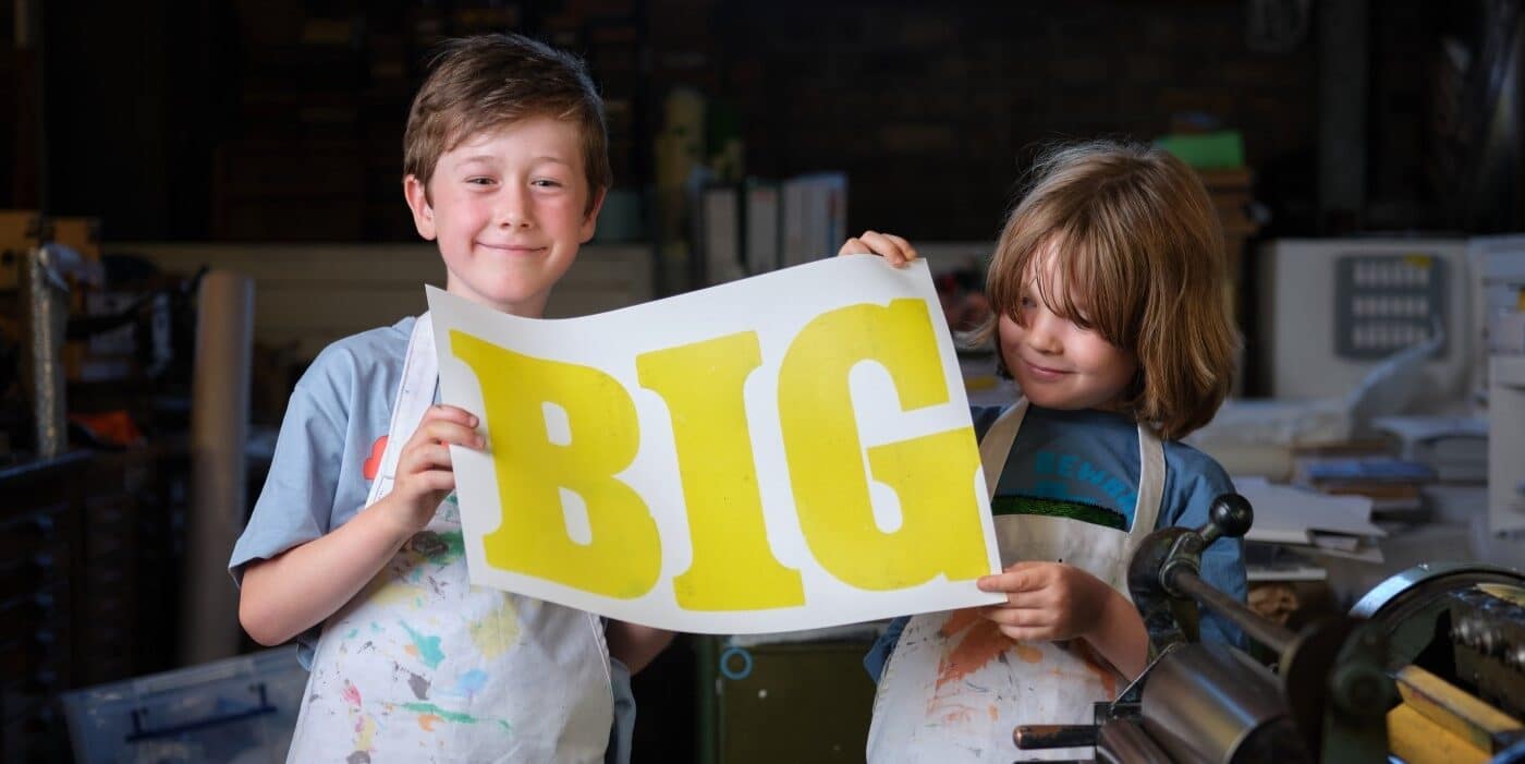 Two young boys hold up a piece of paper with the word BIG printed on it in yellow ink.