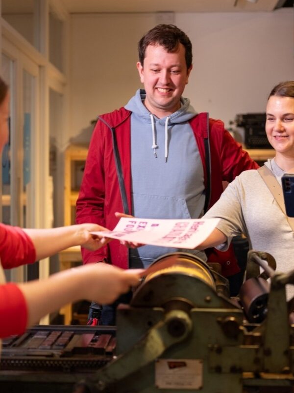 A smiling man and woman receive a print from the hands of a printer beside a printing press.