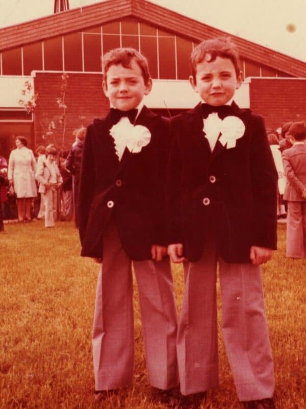 Two young boys in matching clothes stand next to each other in front of a house.