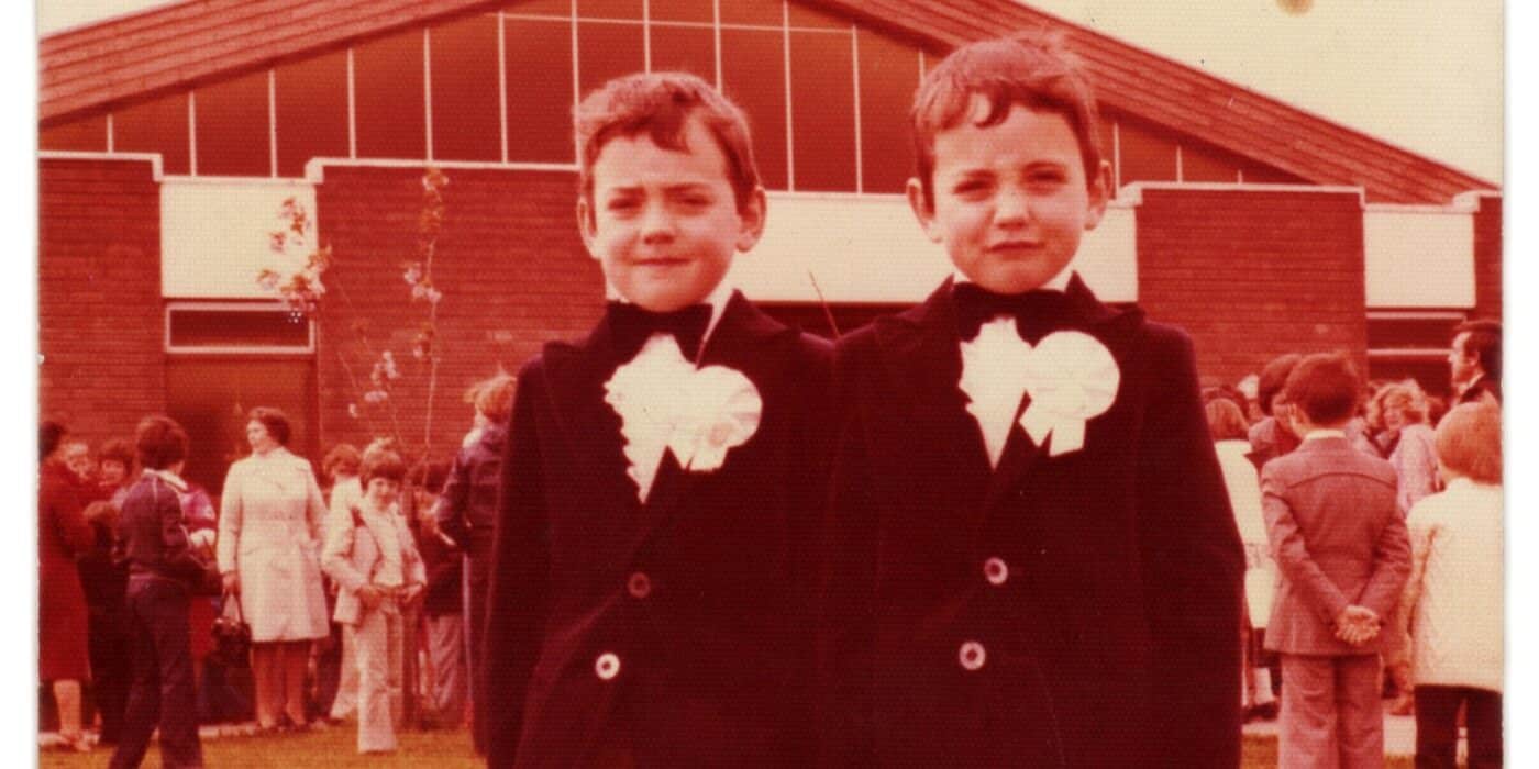 Two young boys in matching clothes stand next to each other in front of a house.