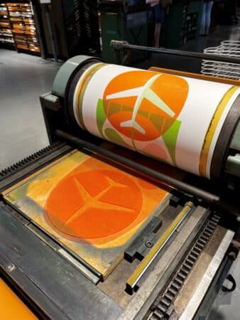 A letterpress print in orange and green of an airplane and a shamrock is in the process of being printed on a printing press.