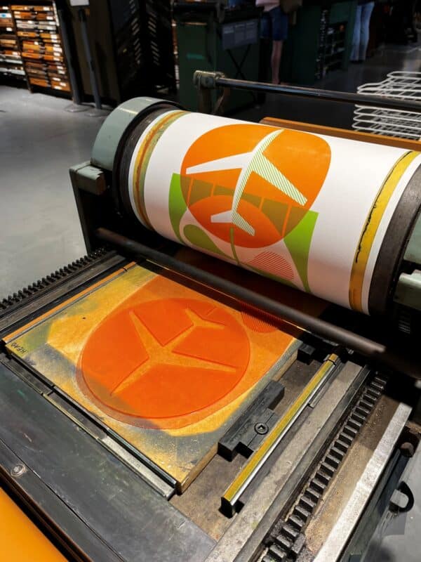 A letterpress print in orange and green of an airplane and a shamrock is in the process of being printed on a printing press.
