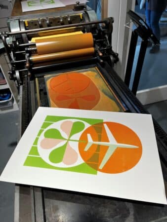 A letterpress print in orange and green of an airplane and a shamrock sits on top of a printing press.